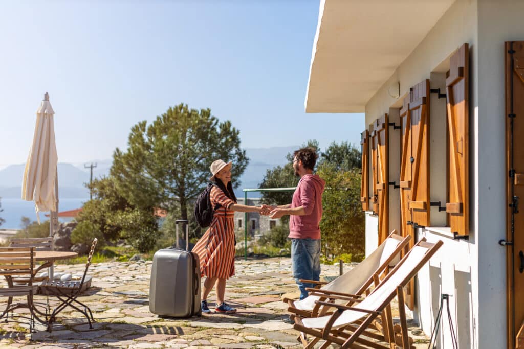 Voyage en couple : arrivée et accueil pour vacances de rêve Couple souriant avec valise se tenant la main sur une terrasse en pierre, devant une maison et la mer. Scène d'arrivée en vacances.