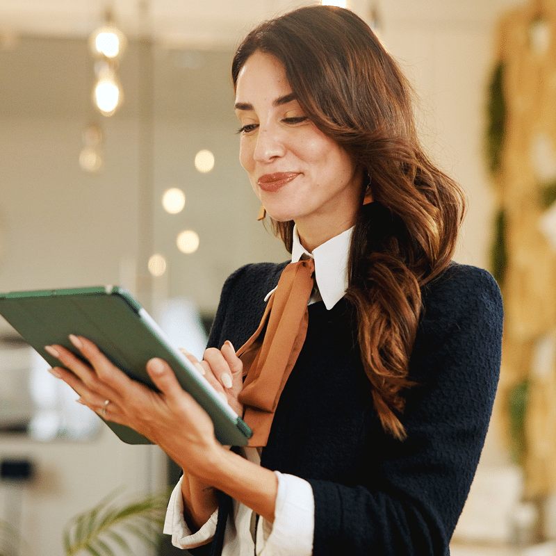 Femme professionnelle souriante naviguant sur une tablette verte. Cheveux bruns, chemise blanche, nœud marron, veste noire.
